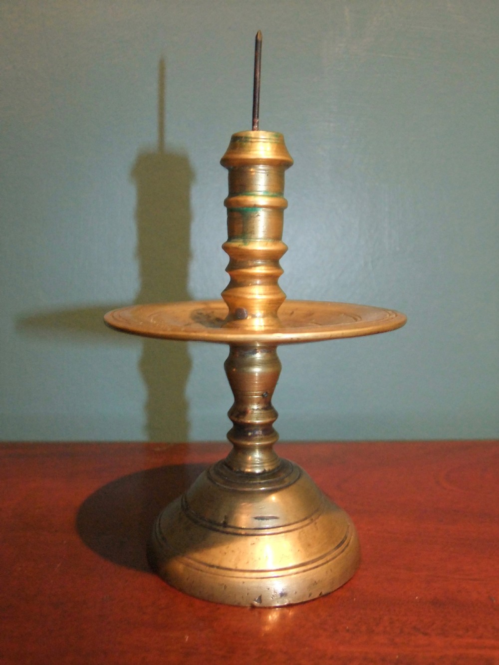 A 17th-century cast brass pricket candlestick featuring a central iron spike, a turned column stem, a wide circular drip tray, and a stepped circular base. View II