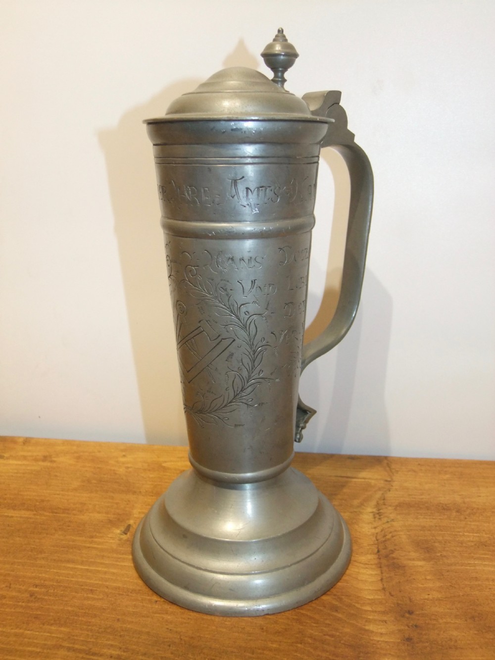 A tall 19th-century German lidded pewter tankard featuring hand-engraved Masonic iconography, floral sprigs, German text, a hinged lid with finial, and a flared pedestal base.