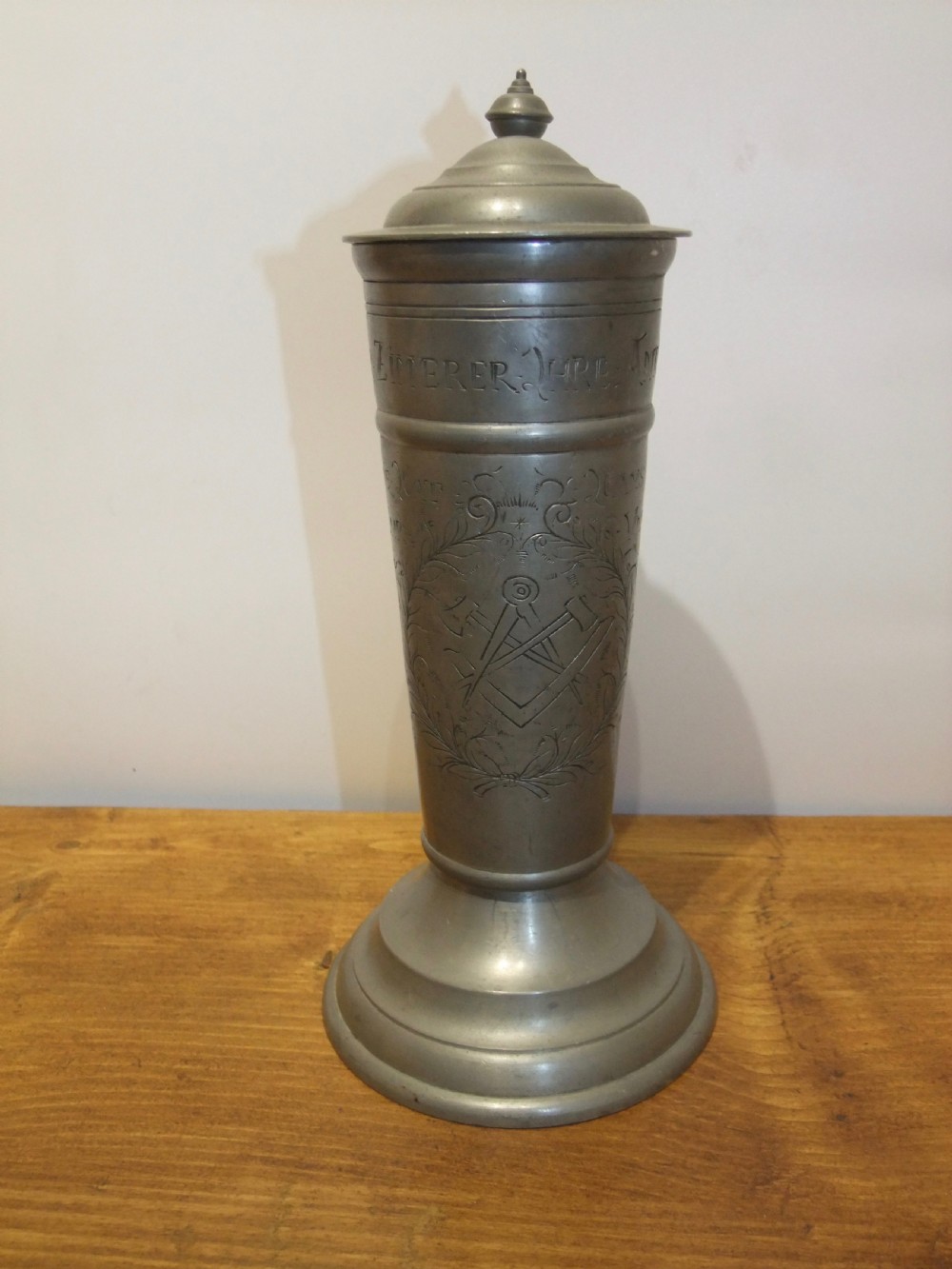 A tall 19th-century German lidded pewter tankard featuring hand-engraved Masonic iconography, floral sprigs, German text, a hinged lid with finial, and a flared pedestal base. View II