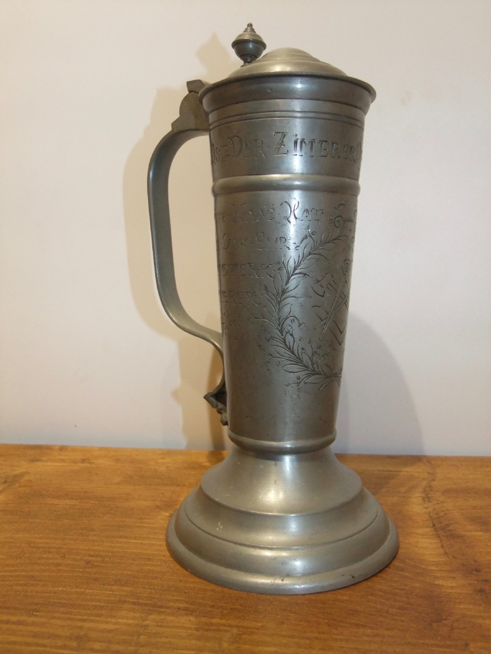 A tall 19th-century German lidded pewter tankard featuring hand-engraved Masonic iconography, floral sprigs, German text, a hinged lid with finial, and a flared pedestal base. View III
