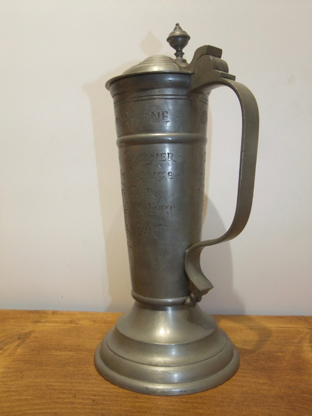 A tall 19th-century German lidded pewter tankard featuring hand-engraved Masonic iconography, floral sprigs, German text, a hinged lid with finial, and a flared pedestal base. View V