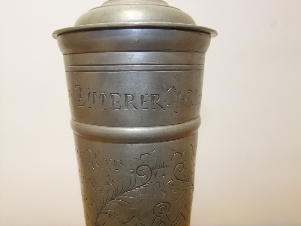 A tall 19th-century German lidded pewter tankard featuring hand-engraved Masonic iconography, floral sprigs, German text, a hinged lid with finial, and a flared pedestal base. View IX