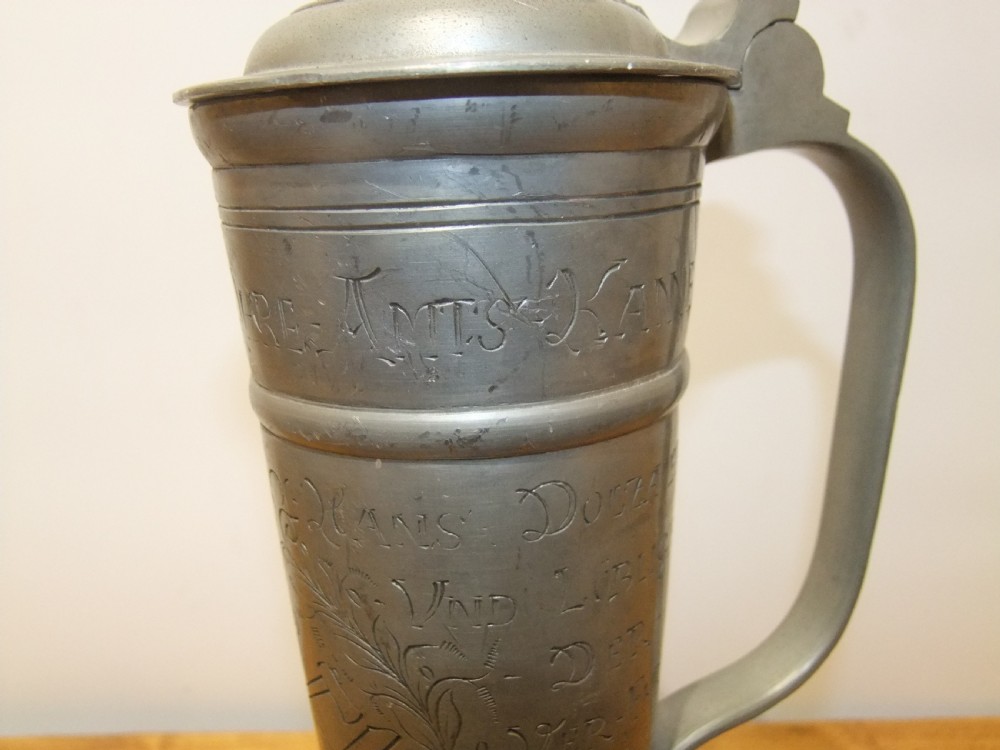 A tall 19th-century German lidded pewter tankard featuring hand-engraved Masonic iconography, floral sprigs, German text, a hinged lid with finial, and a flared pedestal base. View X