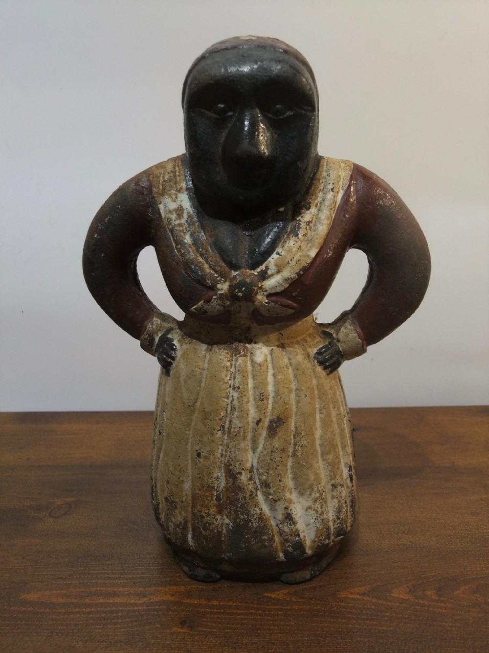 A nineteenth-century painted cast iron money bank depicting a standing figure with hands on hips wearing a red top and cream apron.