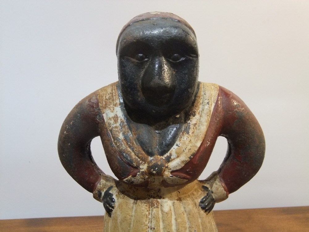 A nineteenth-century painted cast iron money bank depicting a standing figure with hands on hips wearing a red top and cream apron. View II