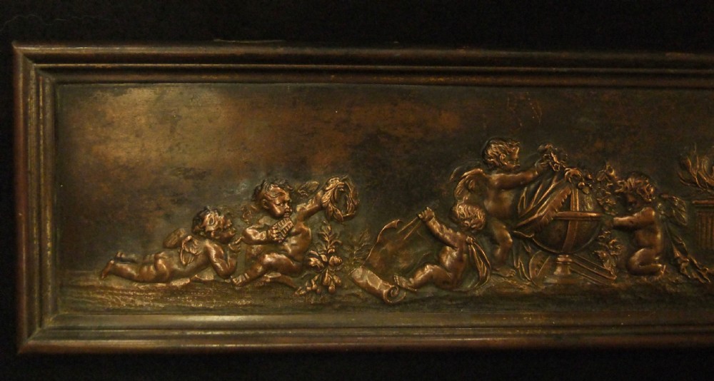 A long horizontal dark bronze relief frieze featuring classical cherubs engaged in allegorical scenes set within a slender rectangular frame. View II