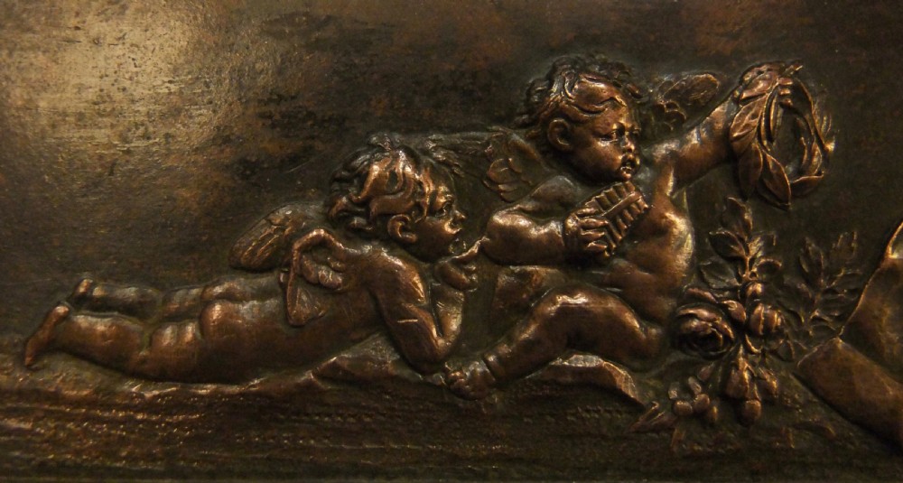 A long horizontal dark bronze relief frieze featuring classical cherubs engaged in allegorical scenes set within a slender rectangular frame. View VI