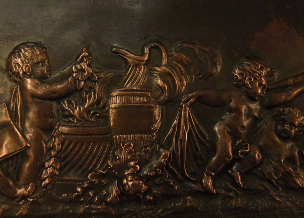 A long horizontal dark bronze relief frieze featuring classical cherubs engaged in allegorical scenes set within a slender rectangular frame. View X