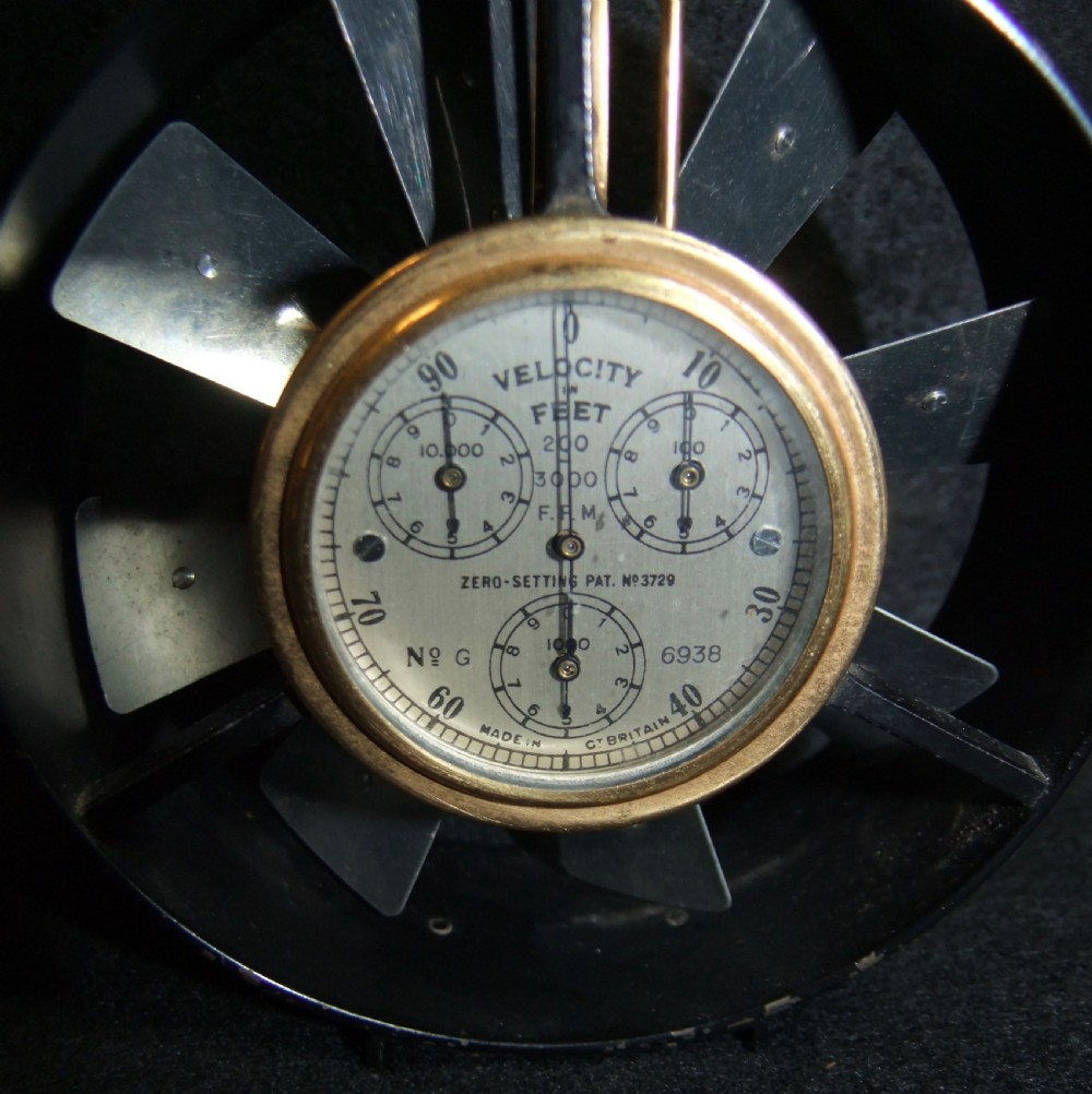 A circular brass and black metal anemometer with fan blades and a multi-dial face, displayed alongside its fitted brown leather carrying case. View IV