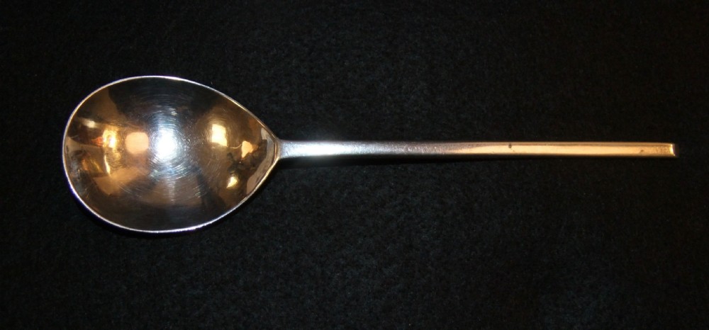 A solid silver spoon featuring a rounded fig-shaped bowl and a straight stem with an obliquely cut slip top end against a dark background.