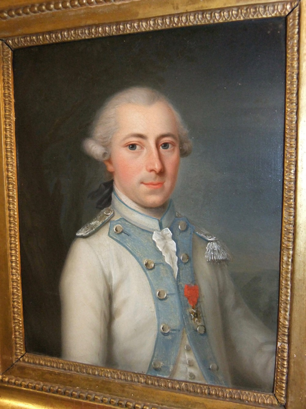 An 18th-century oil portrait depicting a male military officer in a white and blue uniform inside a molded gold gilded frame. View II