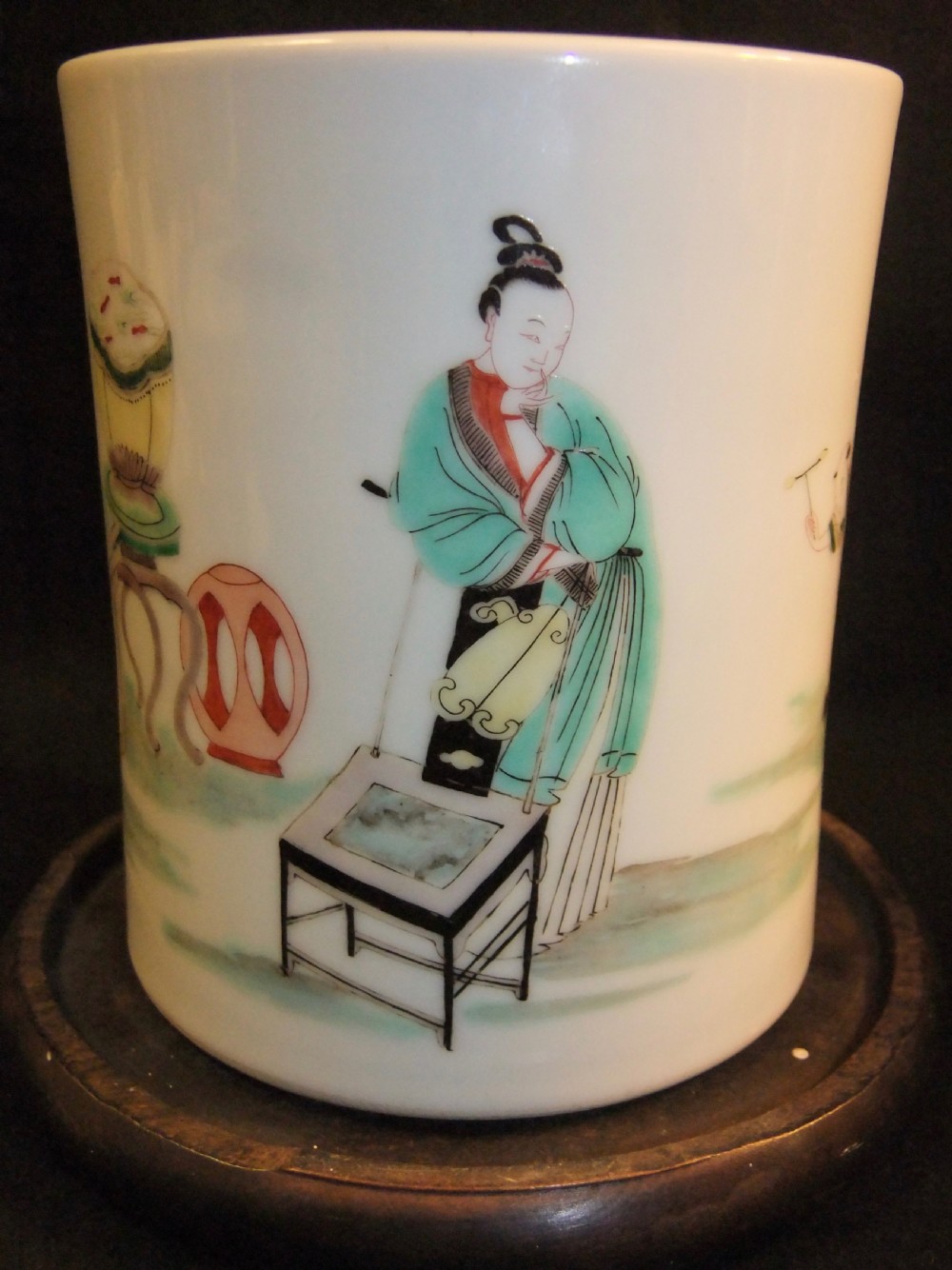 A cylindrical white porcelain brush pot hand-painted with green, red, and yellow enamels depicting two figures, resting on a dark timber stand. View II