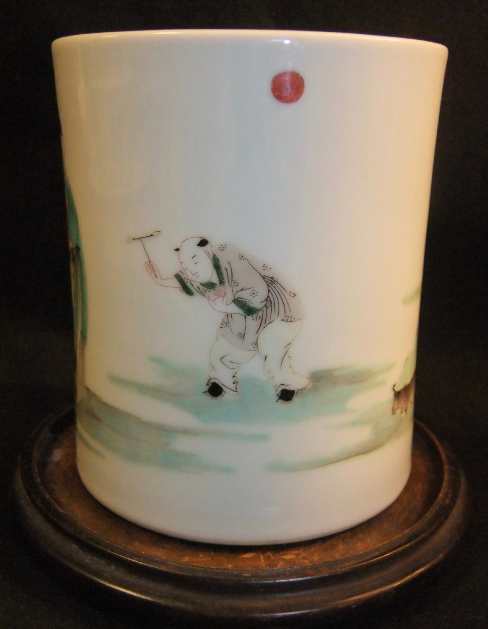 A cylindrical white porcelain brush pot hand-painted with green, red, and yellow enamels depicting two figures, resting on a dark timber stand. View III