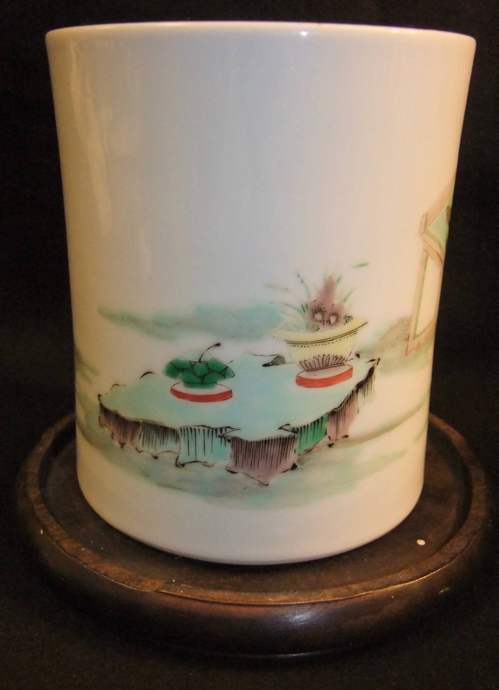 A cylindrical white porcelain brush pot hand-painted with green, red, and yellow enamels depicting two figures, resting on a dark timber stand. View IV