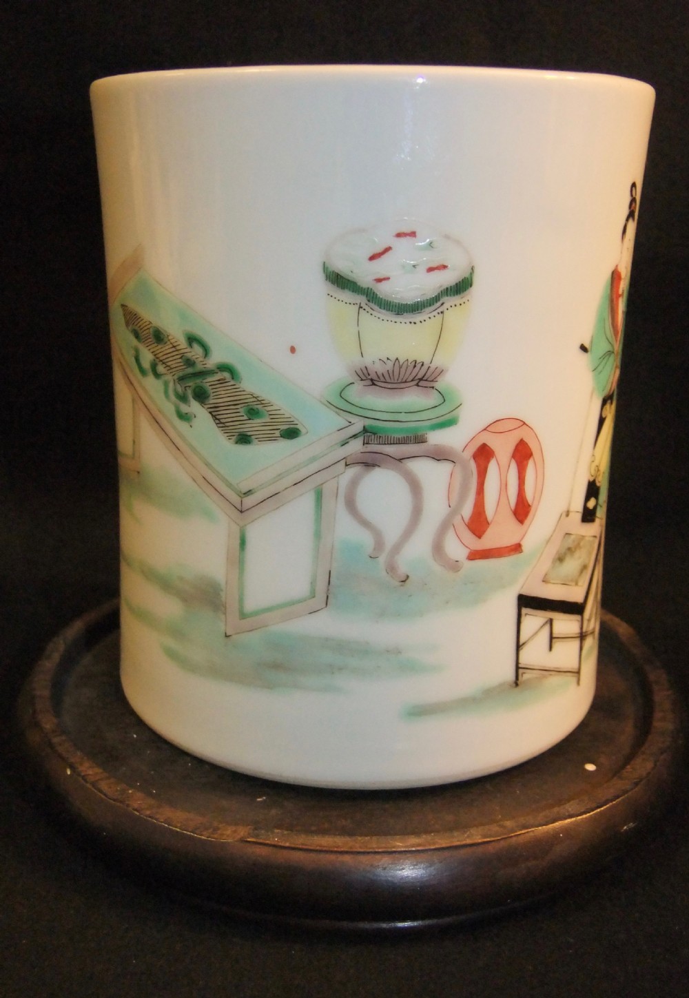 A cylindrical white porcelain brush pot hand-painted with green, red, and yellow enamels depicting two figures, resting on a dark timber stand. View V