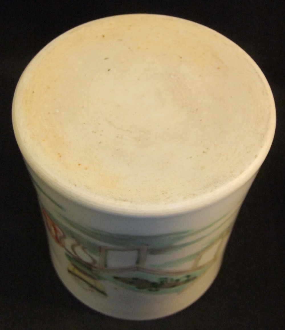 A cylindrical white porcelain brush pot hand-painted with green, red, and yellow enamels depicting two figures, resting on a dark timber stand. View VI