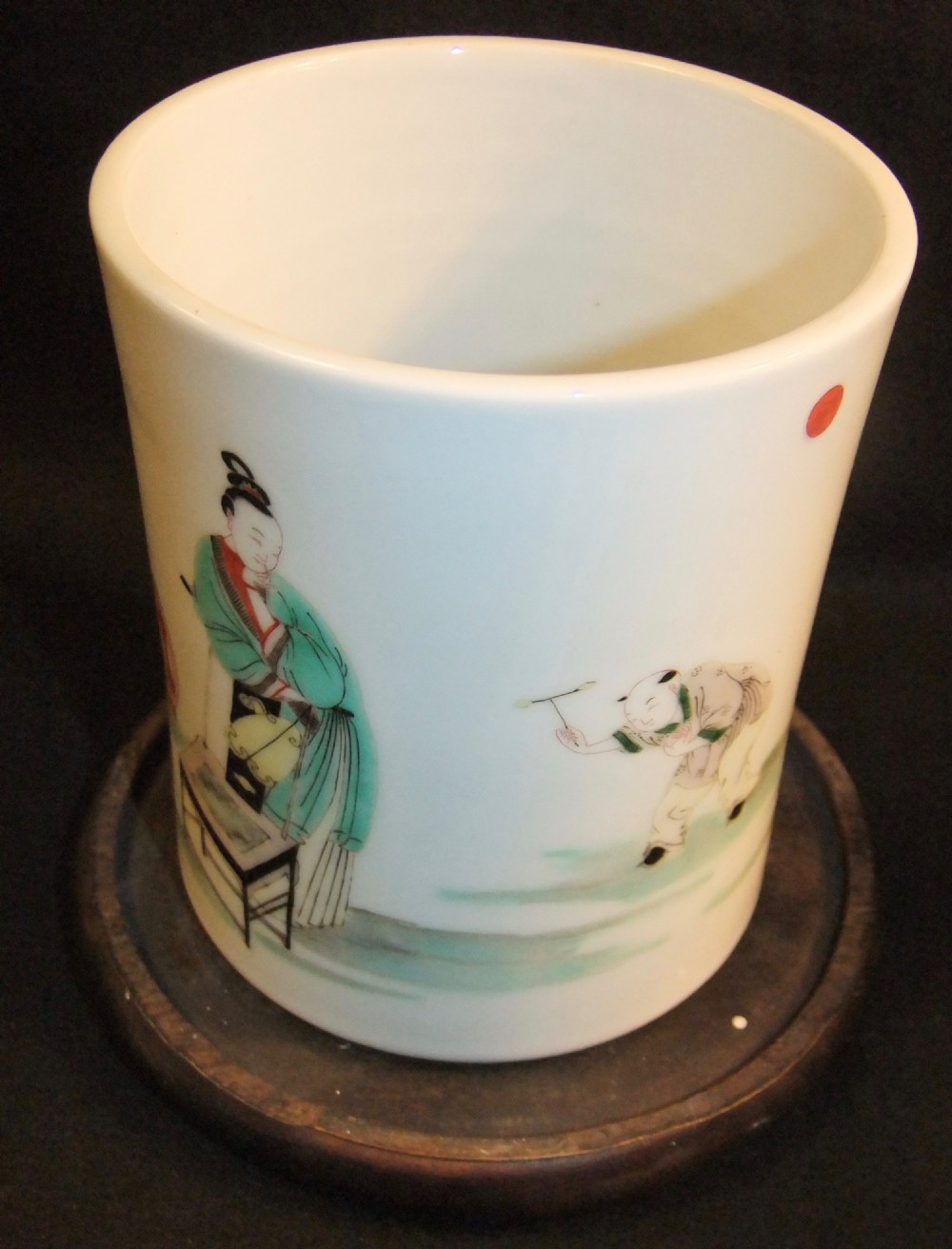 A cylindrical white porcelain brush pot hand-painted with green, red, and yellow enamels depicting two figures, resting on a dark timber stand. View VII