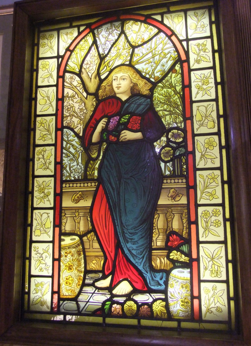 A Pre-Raphaelite stained glass panel featuring a draped female figure surrounded by blossoms and framed by botanical pattern tiles.