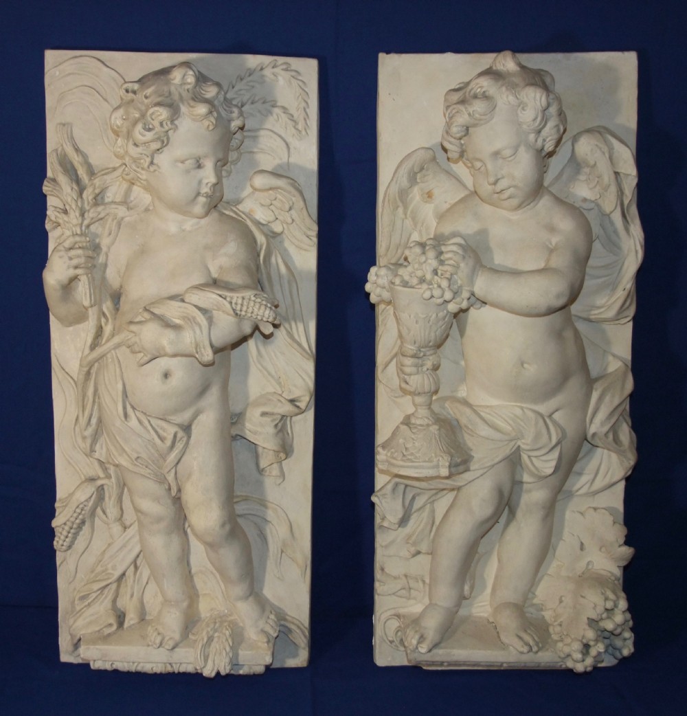 A pair of vertical off-white plaster relief friezes depicting winged cherubs holding wheat sheaves and a chalice with grapes.