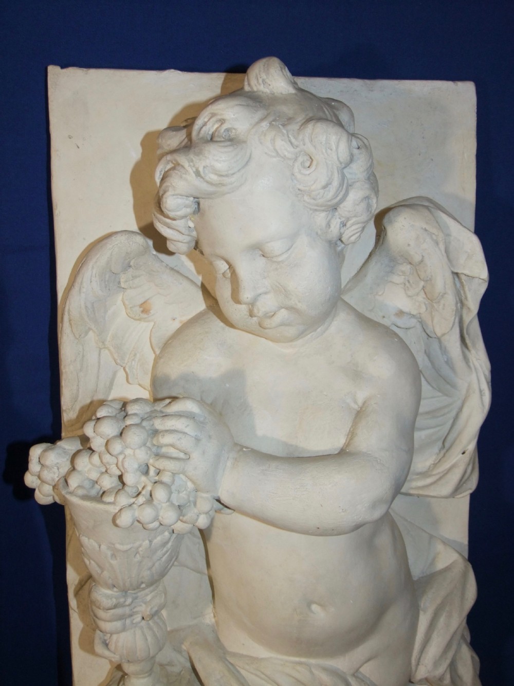 A pair of vertical off-white plaster relief friezes depicting winged cherubs holding wheat sheaves and a chalice with grapes. View V