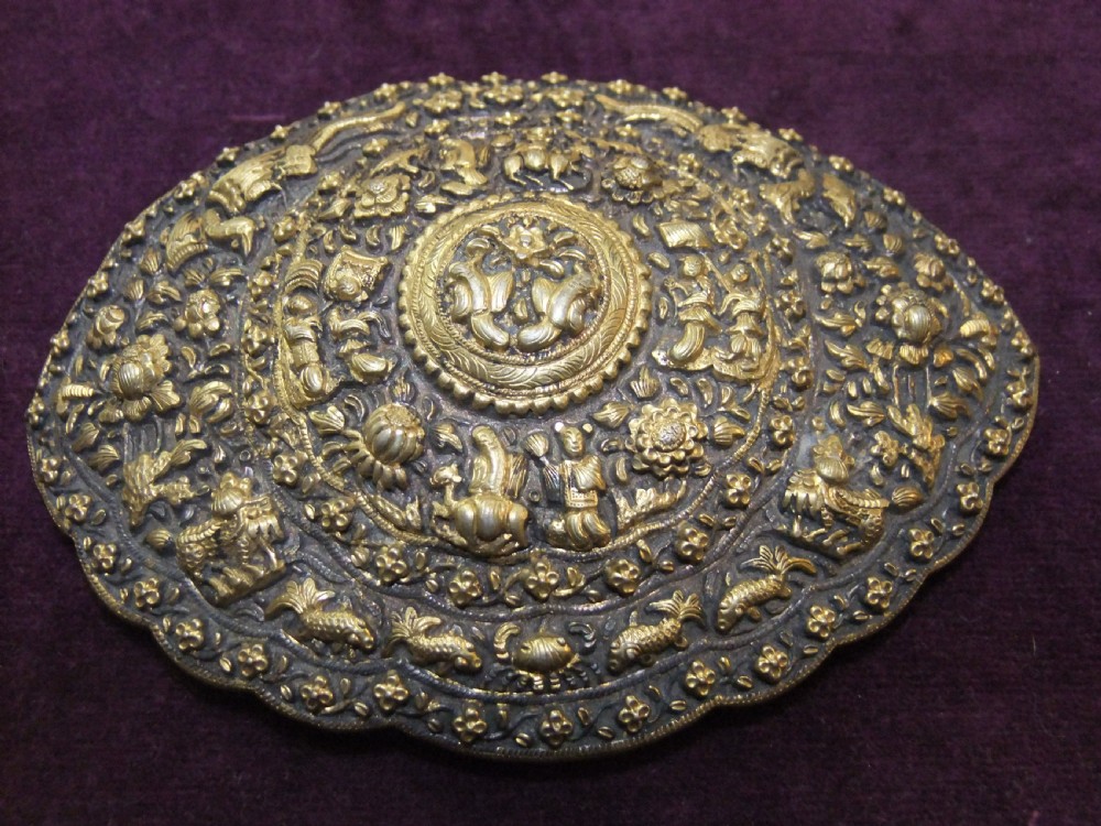 An oval-shaped Straits Chinese gilt silver belt buckle featuring intricate repousse relief carving of figures, floral patterns, and fauna surrounded by a scalloped border.