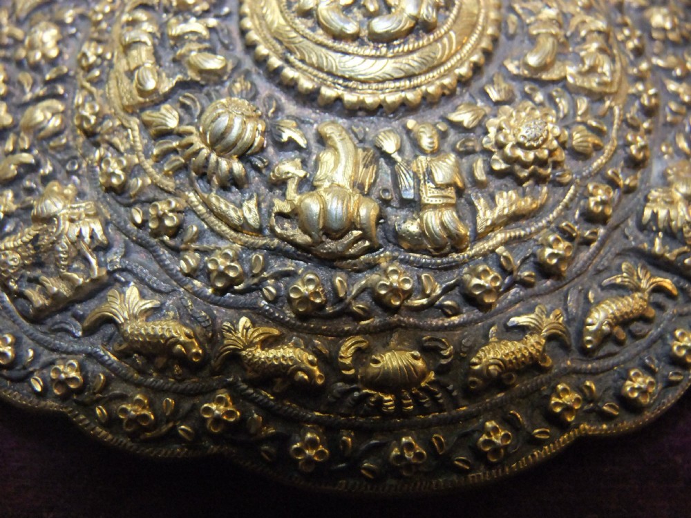 An oval-shaped Straits Chinese gilt silver belt buckle featuring intricate repousse relief carving of figures, floral patterns, and fauna surrounded by a scalloped border. View II