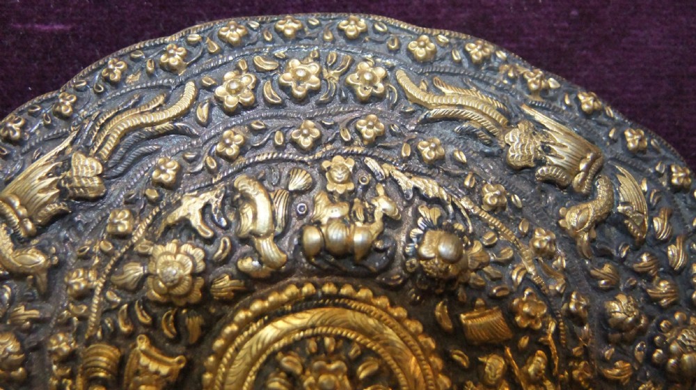 An oval-shaped Straits Chinese gilt silver belt buckle featuring intricate repousse relief carving of figures, floral patterns, and fauna surrounded by a scalloped border. View III