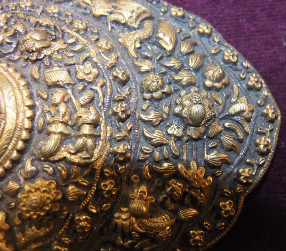 An oval-shaped Straits Chinese gilt silver belt buckle featuring intricate repousse relief carving of figures, floral patterns, and fauna surrounded by a scalloped border. View IV