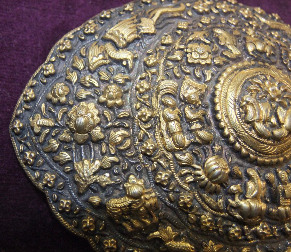 An oval-shaped Straits Chinese gilt silver belt buckle featuring intricate repousse relief carving of figures, floral patterns, and fauna surrounded by a scalloped border. View V