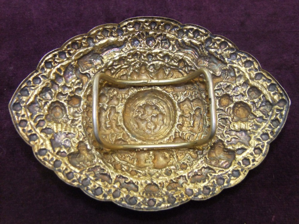 An oval-shaped Straits Chinese gilt silver belt buckle featuring intricate repousse relief carving of figures, floral patterns, and fauna surrounded by a scalloped border. View VI