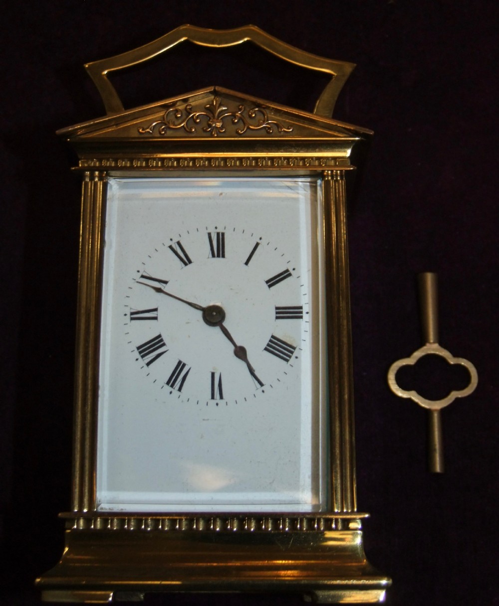 An antique polished brass Georgian revival carriage clock featuring a white dial with Roman numerals, a top carrying handle, and an accompanying winding key.