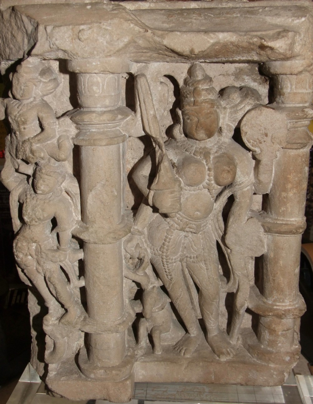 A carved beige sandstone relief fragment from 11th century India depicting the Hindu deity Parvati flanked by decorative columns and smaller attendant figures.