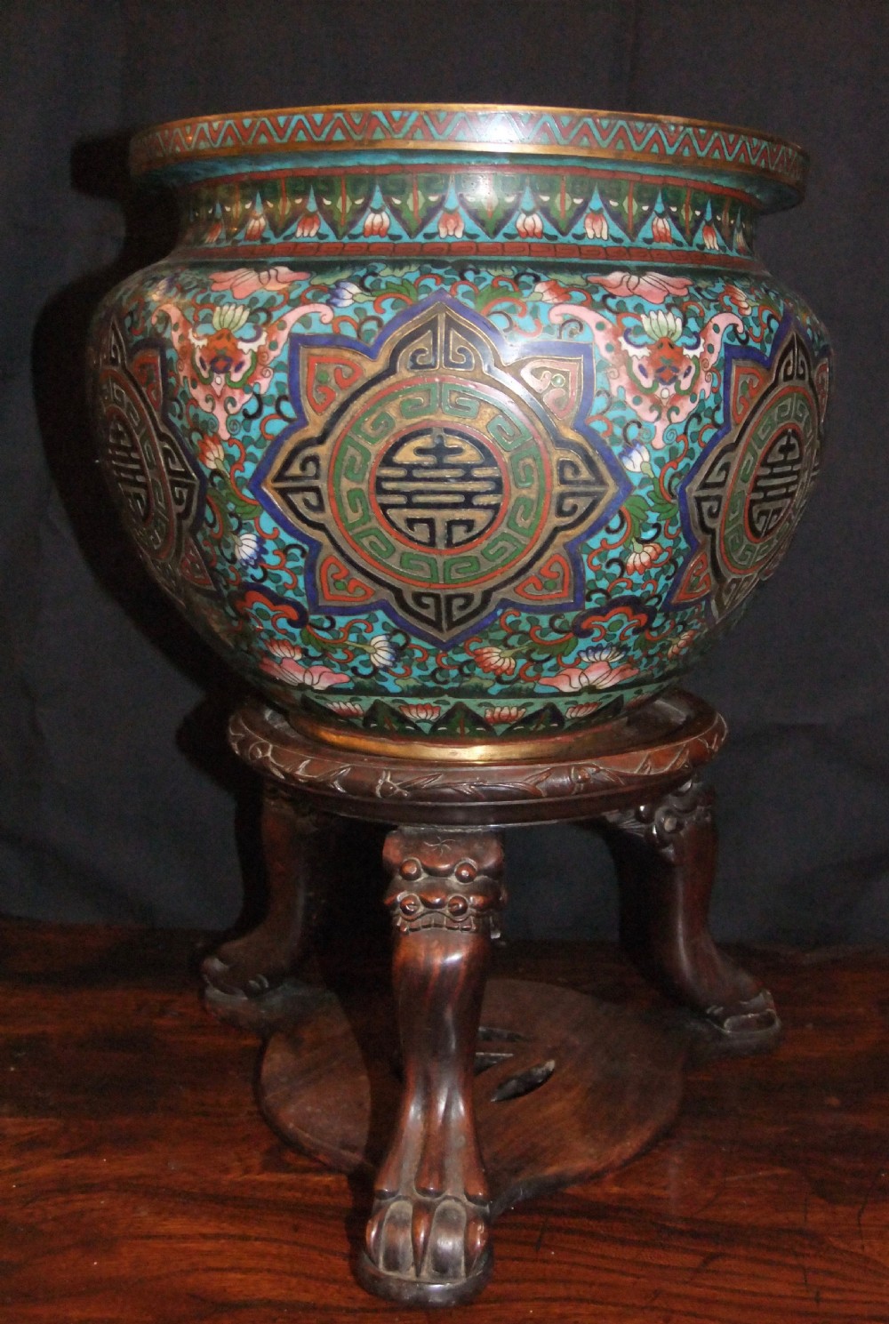 A large Chinese cloisonne enamel jardiniere with floral scrollwork and longevity motifs resting on a dark carved wood stand.