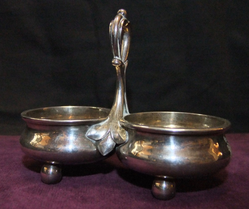 A 19th-century silver double salt cellar featuring twin circular bowls, spherical ball feet, and a central elevated looped handle with leaf detailing.