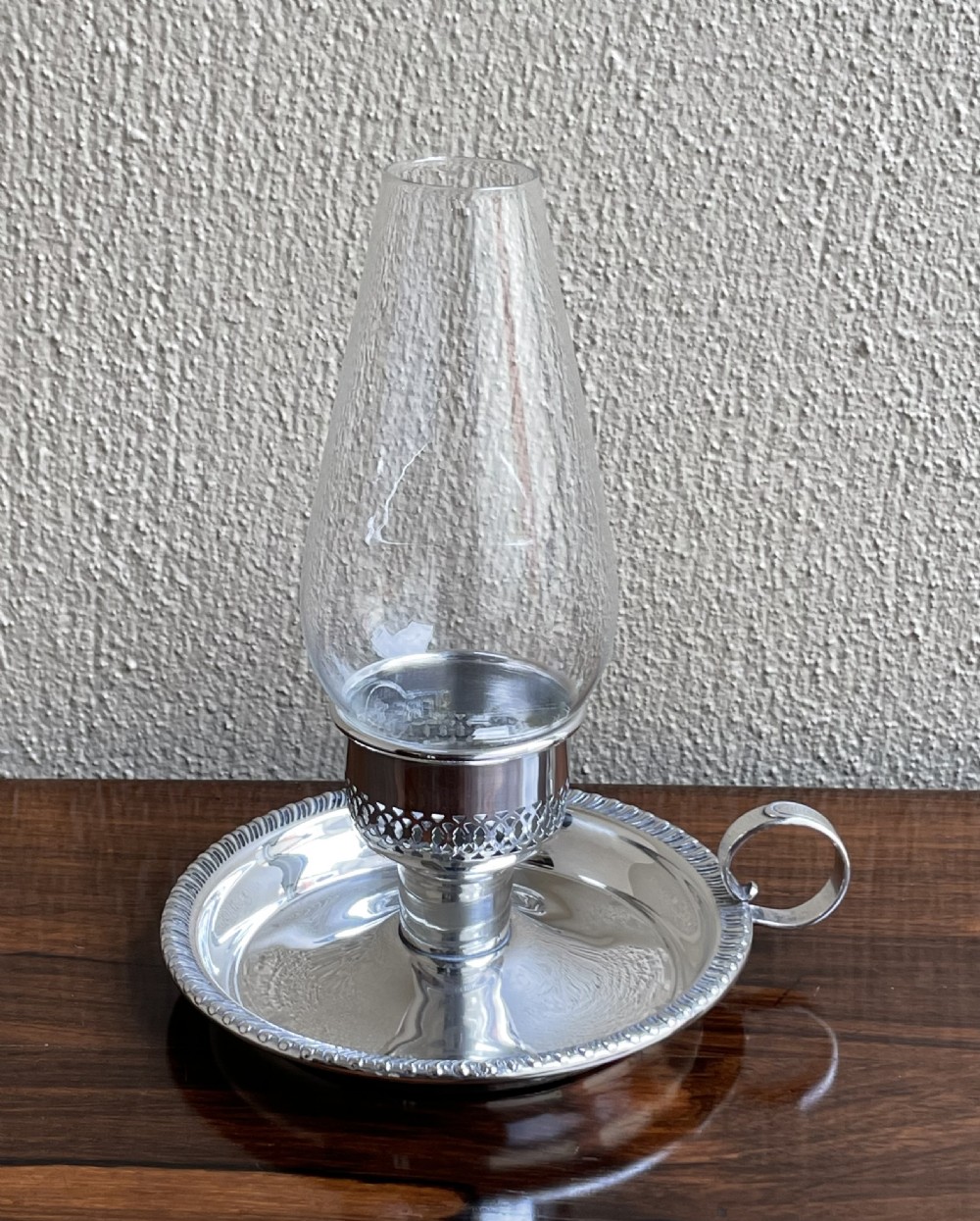 An Art Deco silver chamberstick featuring a tray base with a beaded rim, a pierced candle socket, a side ring handle, and a tall clear glass hurricane shade.
