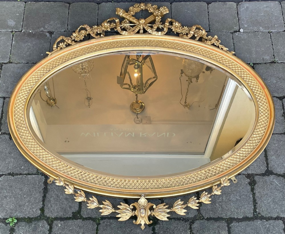 An oval giltwood and gesso wall mirror featuring a ribbon bow and floral garland cresting, a textured diaper pattern frame, and a leaf apron base.