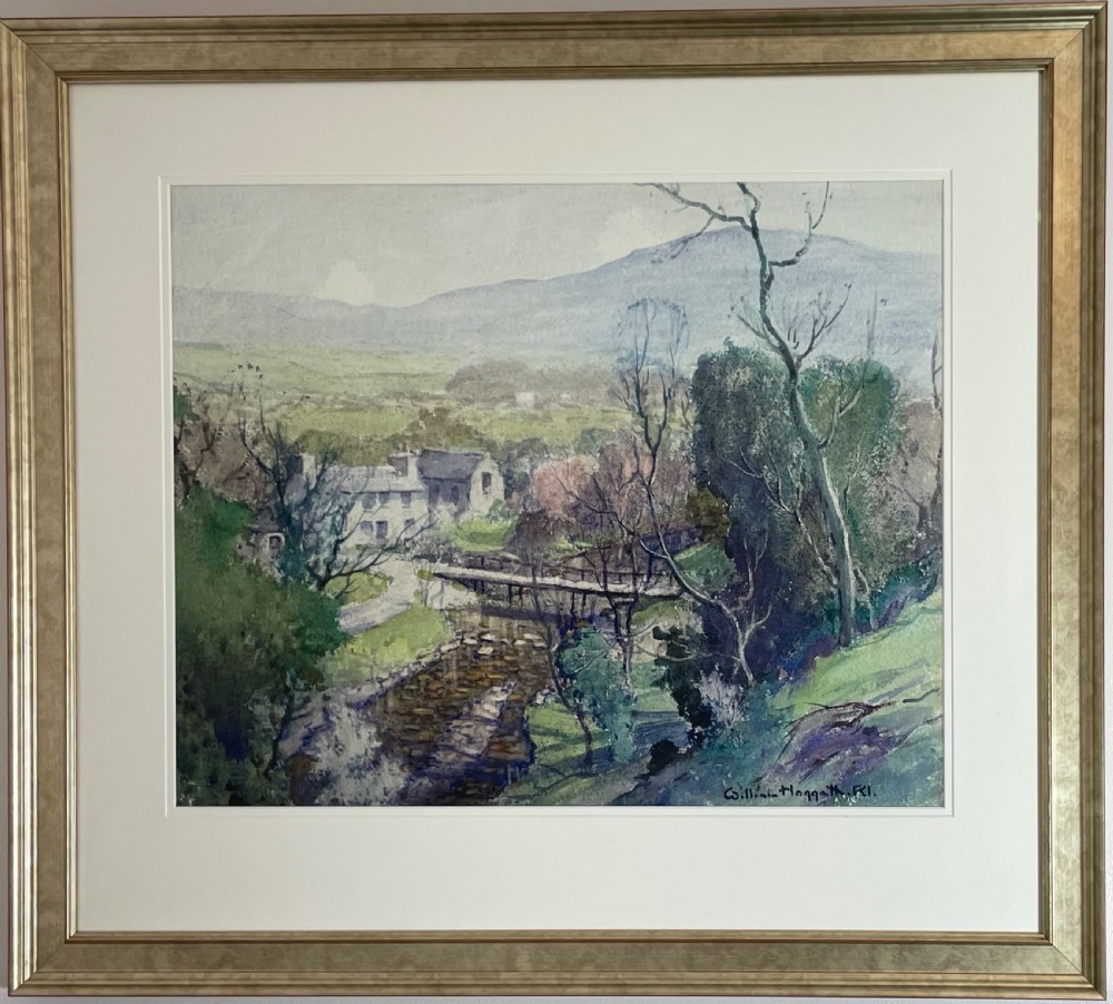 A framed watercolour landscape painting depicting a flowing stream with a wooden footbridge, stone cottages, and distant hills.