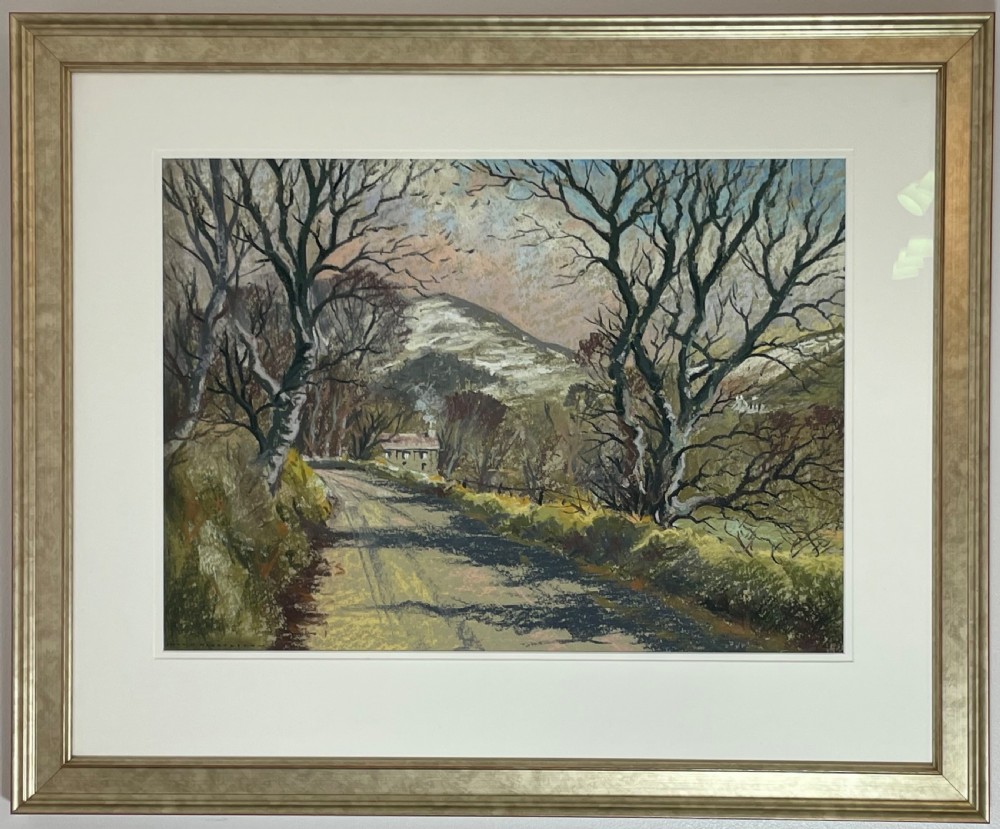 A framed pastel landscape drawing depicting a winding country lane flanked by bare trees with a snow-dusted hill in the background, set in a gold frame with a cream mount.