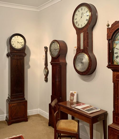 fig 1. An Edwardian dark mahogany longcase clock featuring a circular white dial with Roman numerals, carved ionic capital columns, and a panelled base.