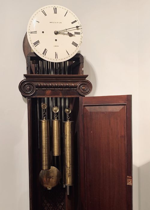 fig 3. An Edwardian dark mahogany longcase clock featuring a circular white dial with Roman numerals, carved ionic capital columns, and a panelled base.
