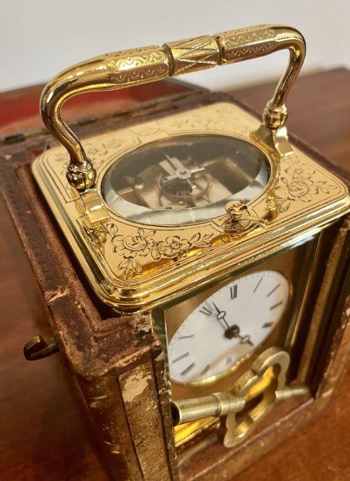 fig 1. A rectangular gilded brass carriage clock featuring an engine-turned faceplate, white enamel dial with Roman numerals, chased floral borders, and a top carrying handle.