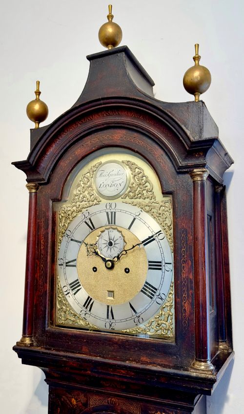 fig 1. A tall antique Georgian Chinoiserie longcase clock with dark gilt lacquered oriental scenes, three brass finials, and a brass dial face.