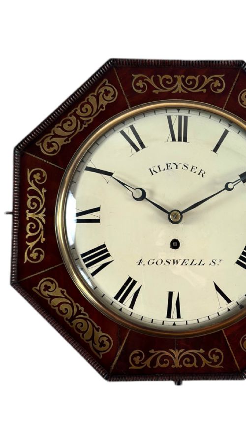 fig 1. An antique octagonal dark wood wall clock featuring scroll brass inlay surrounding a white dial with Roman numerals.
