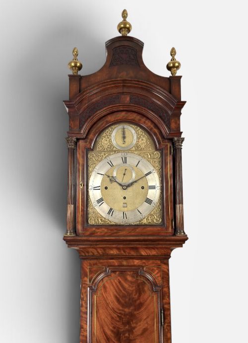 fig 1. An eighteenth-century mahogany longcase clock featuring brass finials, an arched dial, and a figured flame mahogany trunk door.