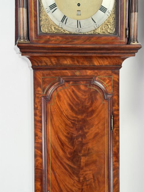 fig 4. An eighteenth-century mahogany longcase clock featuring brass finials, an arched dial, and a figured flame mahogany trunk door.