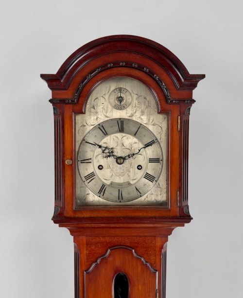 fig 2. A mahogany grandmother clock featuring an arched hood, a silvered clock dial with Roman numerals, and a glazed trunk displaying brass weights.