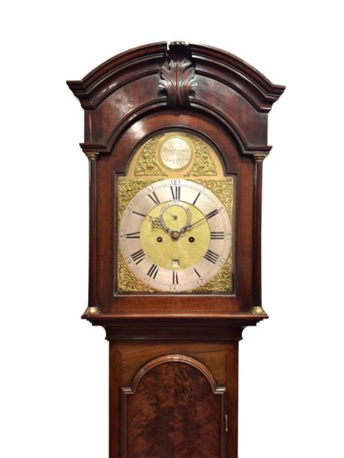 fig 1. A Georgian flame mahogany longcase clock featuring an arched pediment, brass dial with Roman numerals, and a polished dark wood trunk.