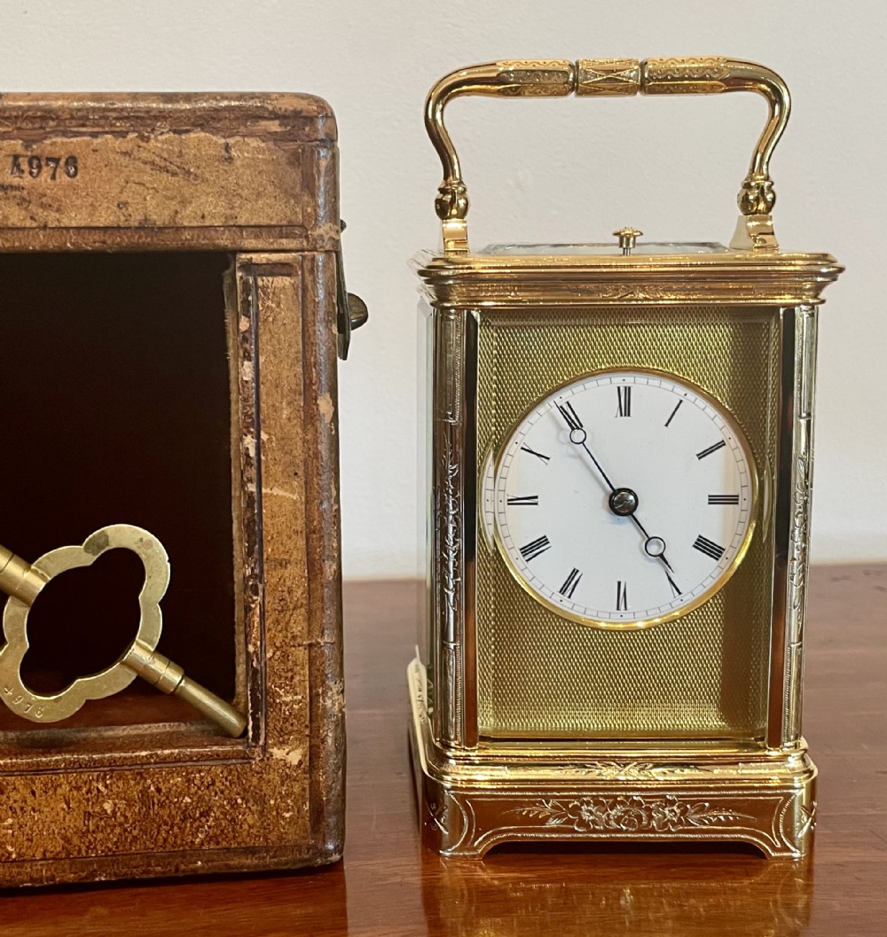 A rectangular gilded brass carriage clock featuring an engine-turned faceplate, white enamel dial with Roman numerals, chased floral borders, and a top carrying handle.