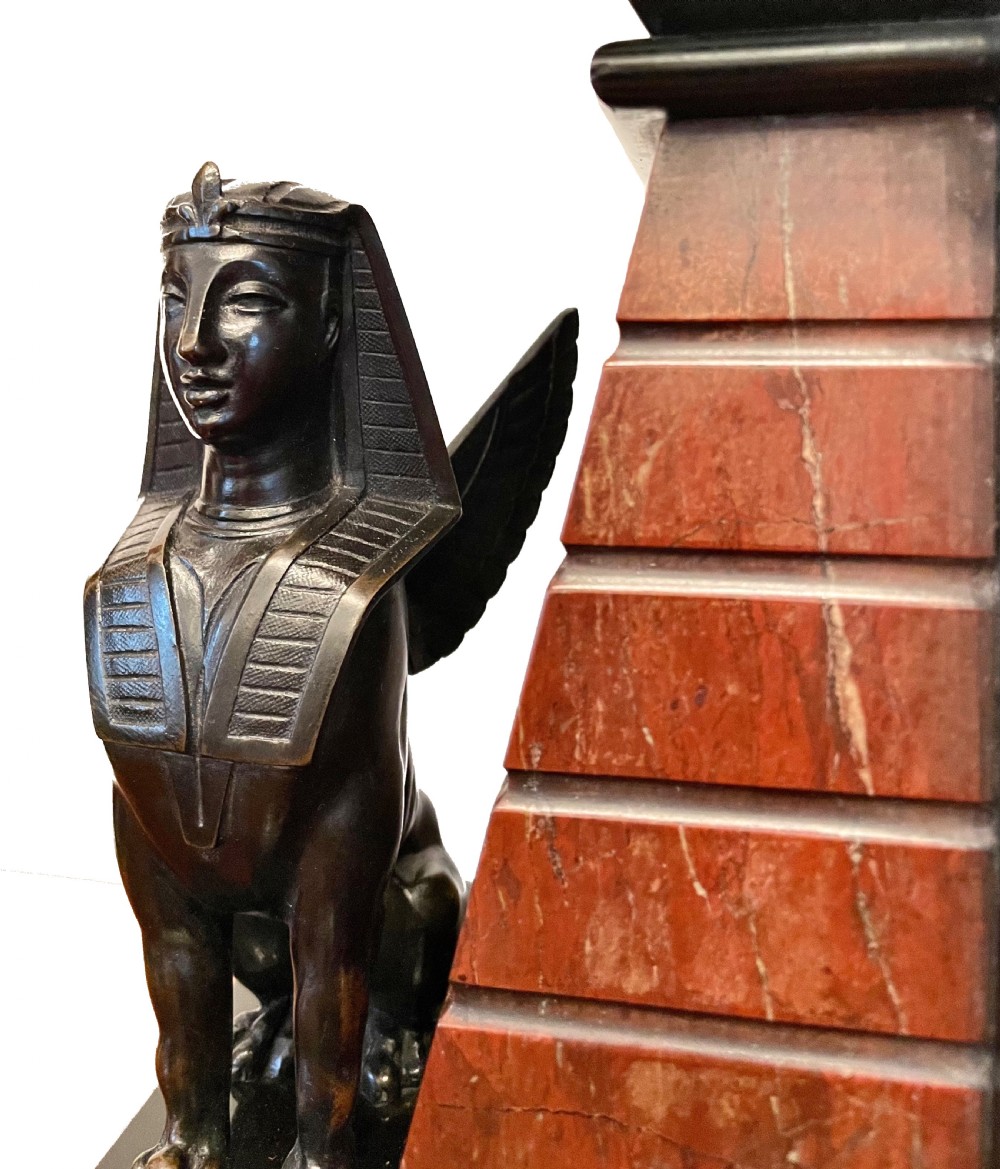 An Egyptian Revival mantel clock constructed from black slate and red marble, topped with a reclining bronze sphinx and flanked by two seated sphinx figures. View II
