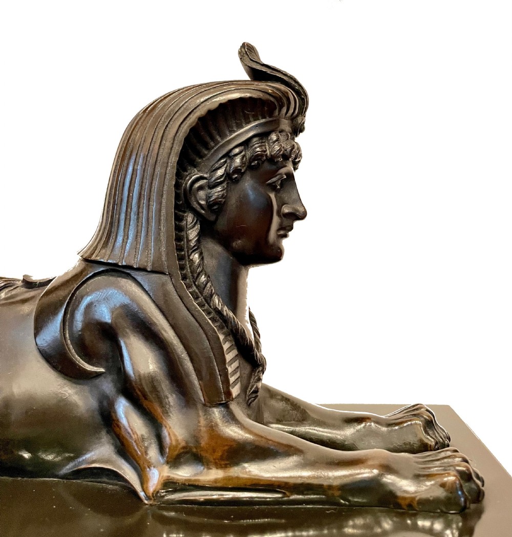 An Egyptian Revival mantel clock constructed from black slate and red marble, topped with a reclining bronze sphinx and flanked by two seated sphinx figures. View III